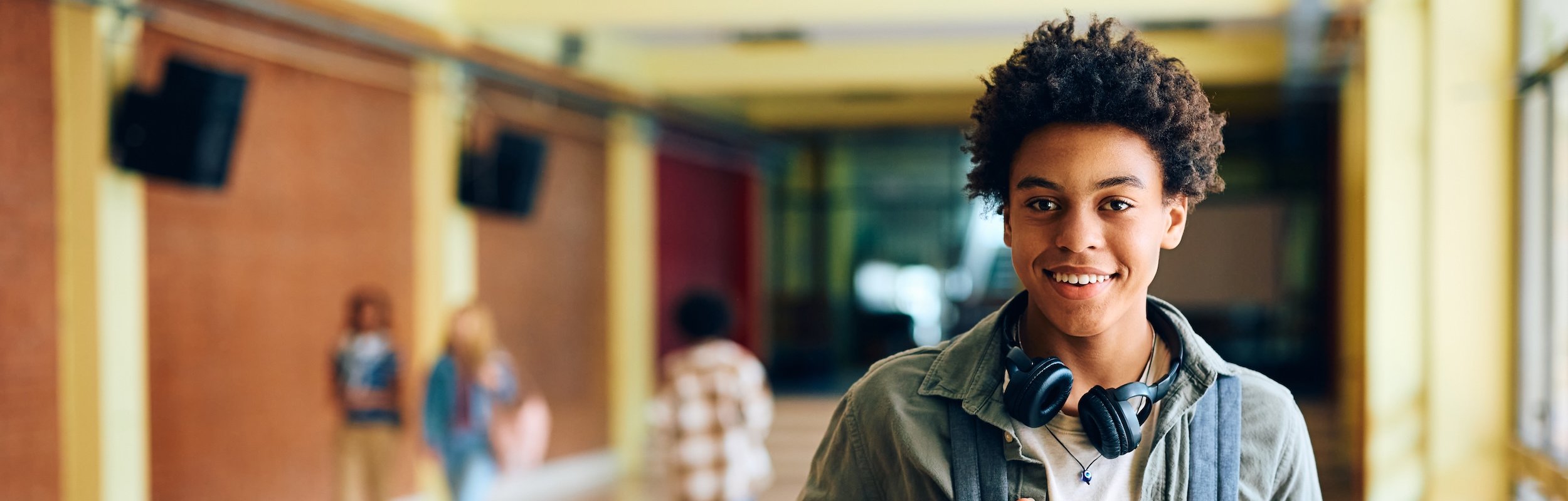 A smiling teenage student walks through a school hallway wearing a backpack and headphones around his neck, with other students visible in the blurred background.